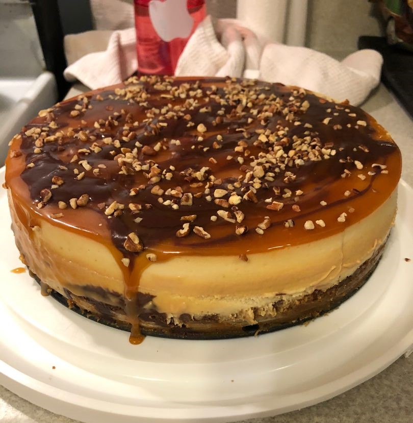 A Pecan Pie Cheesecake with caramel topping and scattered pecans, served on a white plate.